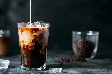 glass with cold brew coffee with cream being poured in coffee beans in the backgroud on table and in glass